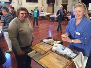 Two women in a hall standing at a table with open boxes. Other people are in the background.