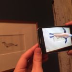Hands holding a smart phone in front of an artwork with an animated seal appearing on the phone.