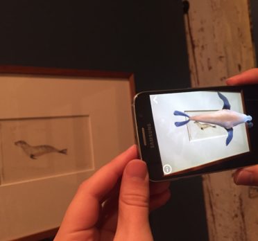 Hands holding a smart phone in front of an artwork with an animated seal appearing on the phone.