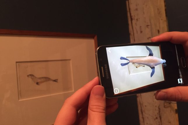 Hands holding a smart phone in front of an artwork with an animated seal appearing on the phone.