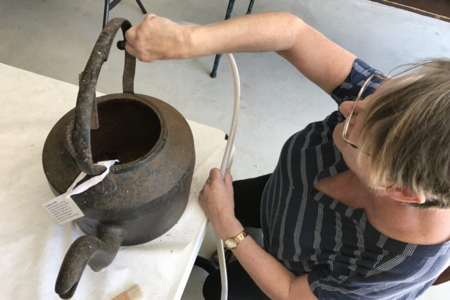 A woman with a vacuum cleaner brush attachment cleaning an old kettle.